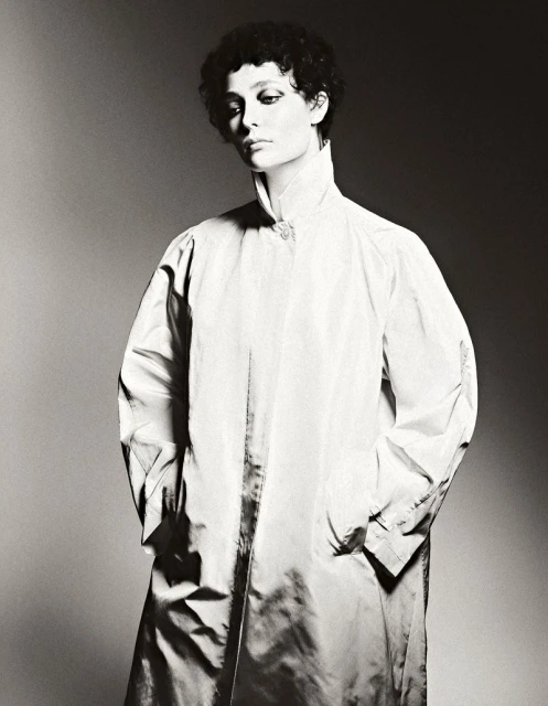Monochrome portrait of a woman with short curly hair wearing a white, high-collared coat, set against a gradient grey background.
