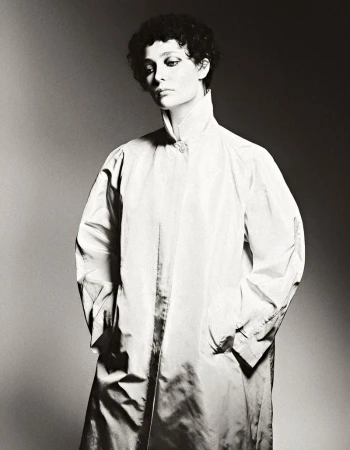 Monochrome portrait of a woman with short curly hair wearing a white, high-collared coat, set against a gradient grey background.