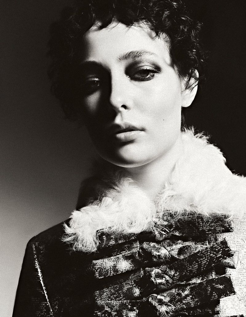 Portrait of an adult woman with curly short hair and dramatic eye makeup wearing a textured jacket with fur collar, black and white.