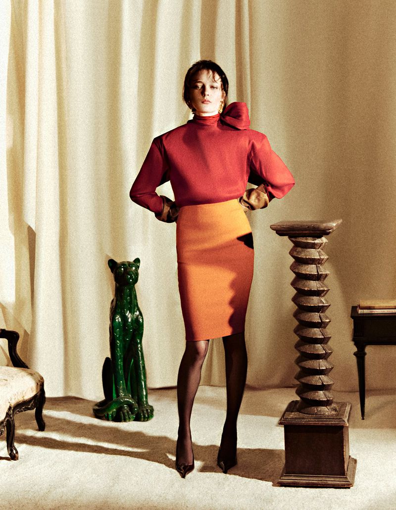 A woman poses in a red top and orange skirt in a vintage-style studio with a ceramic panther, vintage furniture, and neutral backdrop.