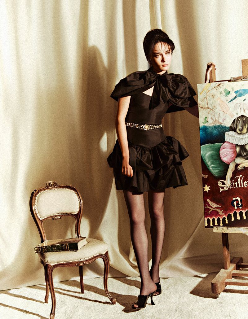Fashion shot of a woman in a black dress standing next to an easel with a dog painting, posing in front of a beige curtain backdrop.