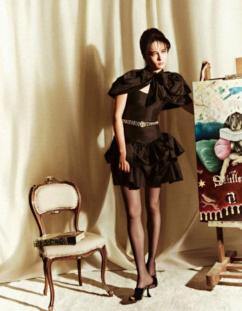 Fashion shot of a woman in a black dress standing next to an easel with a dog painting, posing in front of a beige curtain backdrop.