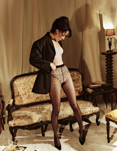 A woman in a blazer, top, and patterned tights poses in a room with vintage furniture, conveying a vintage fashion editorial concept.