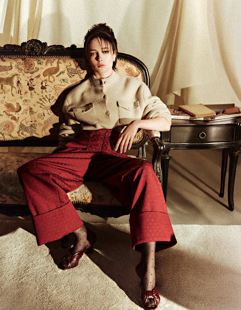 Woman in red pants and cream jacket sits on an ornate chair near a side table in a softly lit interior, fashion editorial.