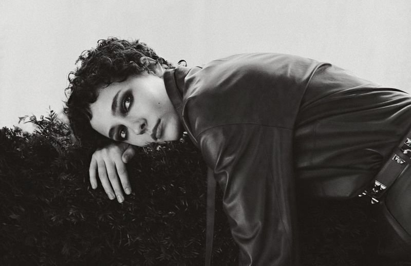 Monochrome portrait of a young adult with dark curly hair, lying on greenery, wearing a black leather jacket with metallic accents.