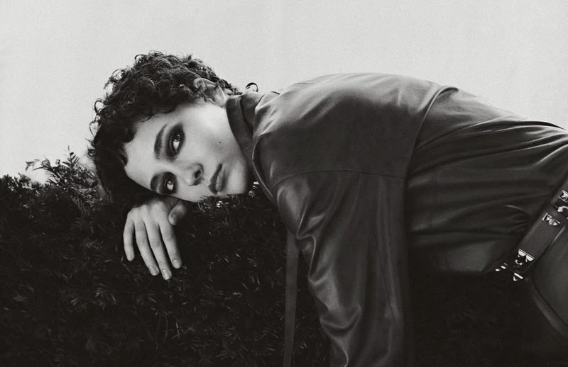 Monochrome portrait of a young adult with dark curly hair, lying on greenery, wearing a black leather jacket with metallic accents.