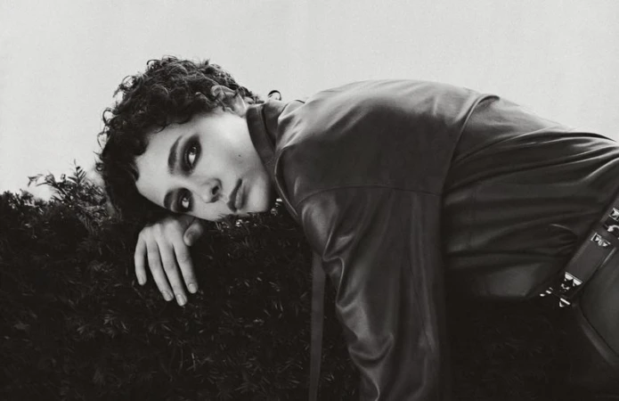 Monochrome portrait of a young adult with dark curly hair, lying on greenery, wearing a black leather jacket with metallic accents.