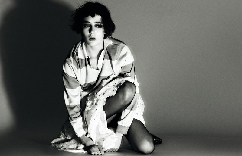 Striking black and white portrait of a young woman kneeling with bare feet, wearing a striped shirt and lacy dress in a studio setting.