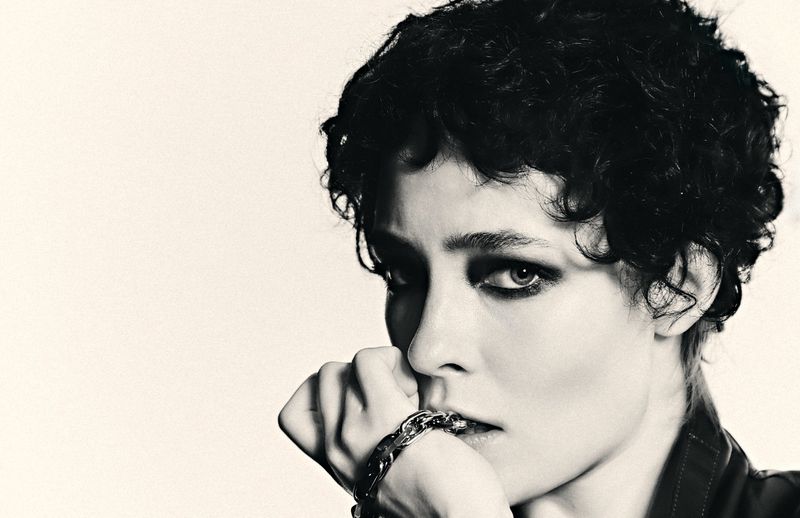 Close-up portrait of a woman with dark curly hair, dramatic eye makeup, and a chain bracelet held to her mouth in black and white.