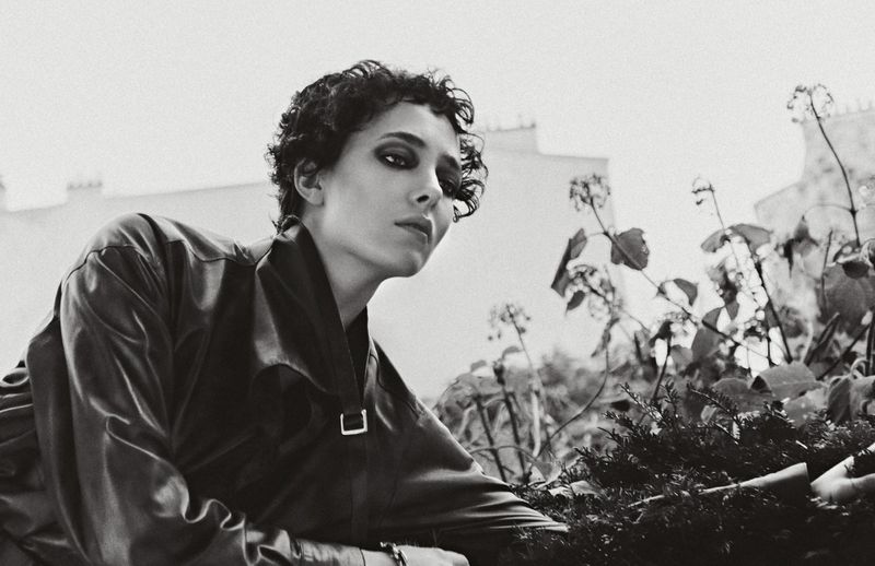 Black-and-white portrait of a woman with short curly hair wearing a jacket and posing outdoors with foliage and a building in the background.