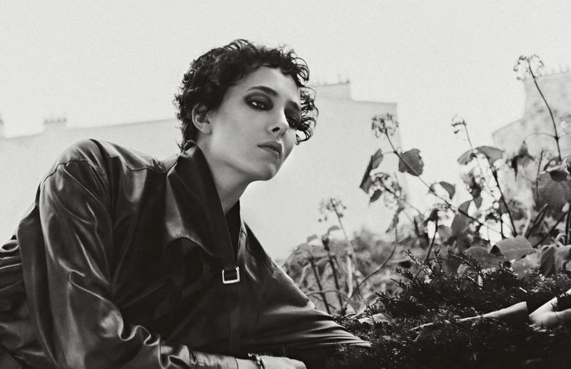 Black-and-white portrait of a woman with short curly hair wearing a jacket and posing outdoors with foliage and a building in the background.