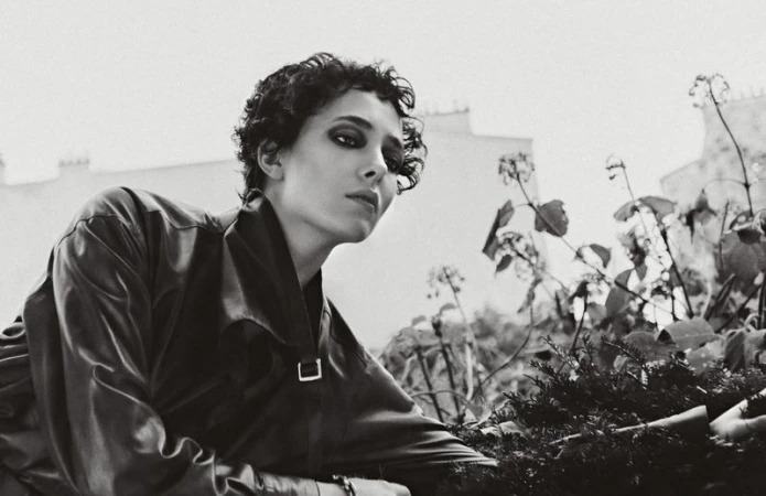 Black-and-white portrait of a woman with short curly hair wearing a jacket and posing outdoors with foliage and a building in the background.