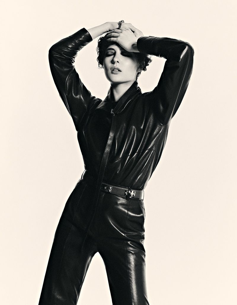 Monochrome portrait of a stylish woman in a black leather jumpsuit, standing in a bright studio with her hands gently placed on her head.