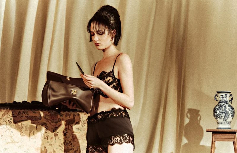 Woman in lingerie with a handbag indoors, posing in front of beige curtains and a vase on a stool, under warm lighting.