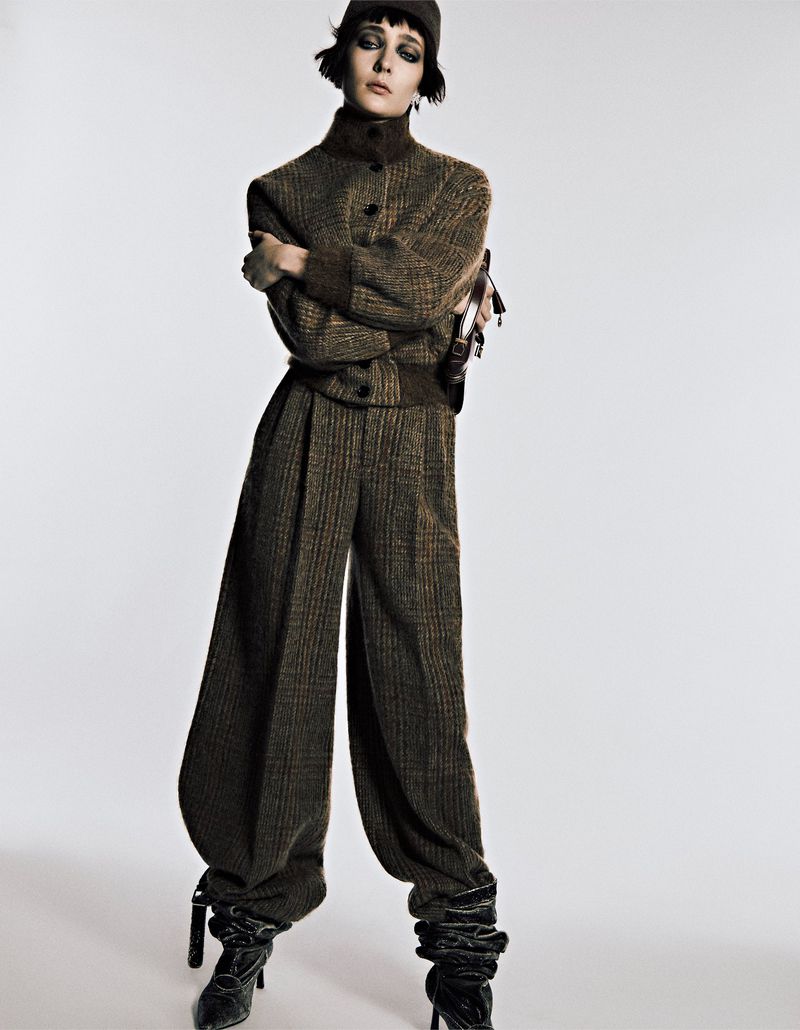Fashion model poses in a studio wearing a brown tweed two-piece suit with balloon pants, black boots, and a cap against a white backdrop.