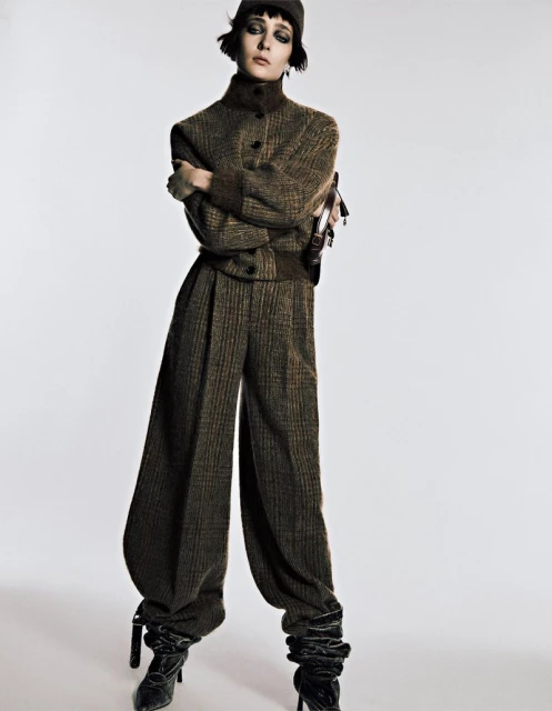 Fashion model poses in a studio wearing a brown tweed two-piece suit with balloon pants, black boots, and a cap against a white backdrop.