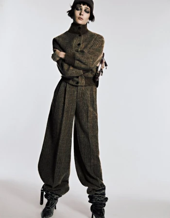 Fashion model poses in a studio wearing a brown tweed two-piece suit with balloon pants, black boots, and a cap against a white backdrop.