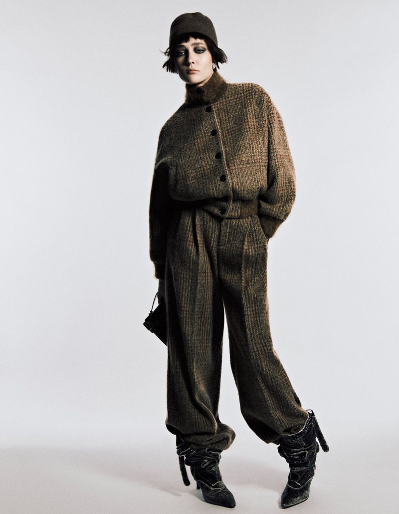 A fashionable adult female model posing in a brown, patterned jacket and pants with boots and a hat against a white backdrop in a photo studio.