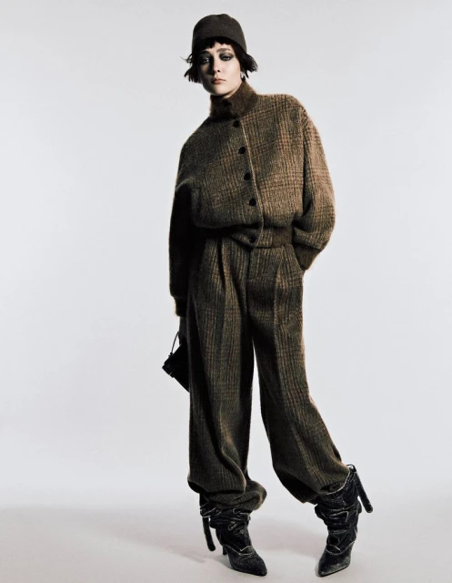 A fashionable adult female model posing in a brown, patterned jacket and pants with boots and a hat against a white backdrop in a photo studio.