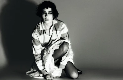 Celeste Fitzpatrick crouching in a striped polo shirt, lace skirt, and black tights against a grey studio backdrop.