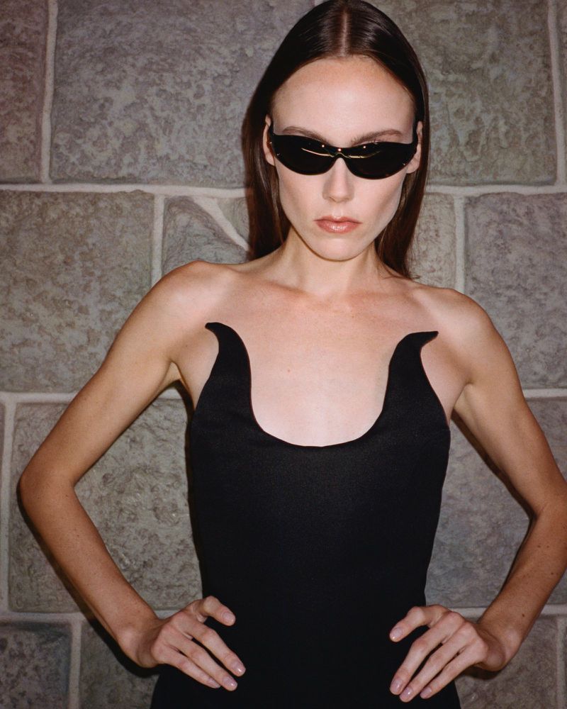 Woman in black dress and sunglasses poses against a gray brick wall indoors. The dress has a unique horn-shaped neckline.