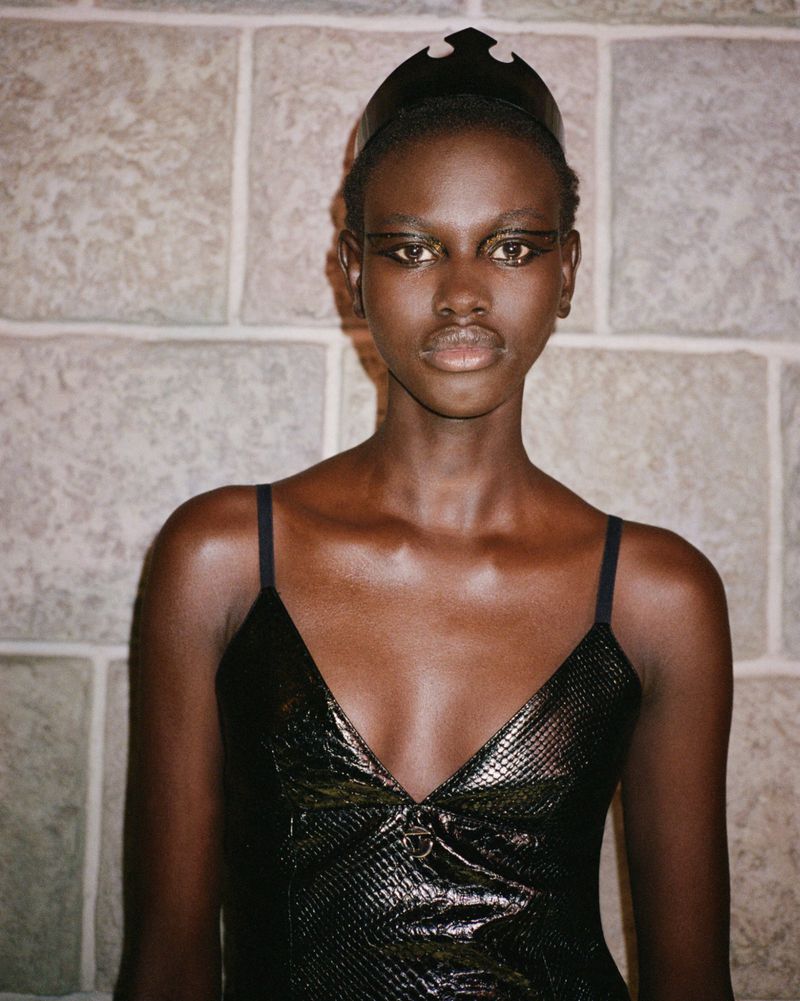 Close-up studio shot of a Black female fashion model with sleek black eyeliner and an avant-garde black headdress, wearing a shiny black top.