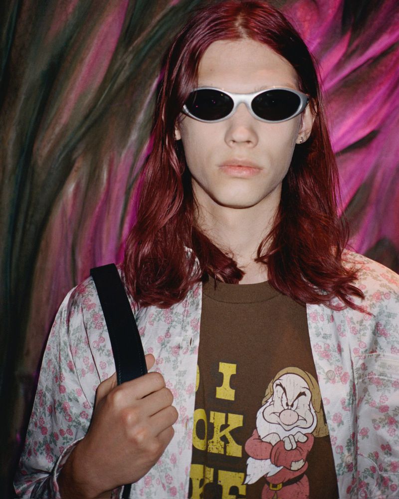 Young adult male with long red hair and sunglasses, wearing a brown T-shirt with a cartoon graphic, in front of a colorful patterned backdrop.