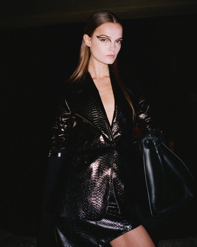 A woman poses in a shiny black 'snakeskin' jacket dress with graphic eyeliner in a studio setting against a dark background.