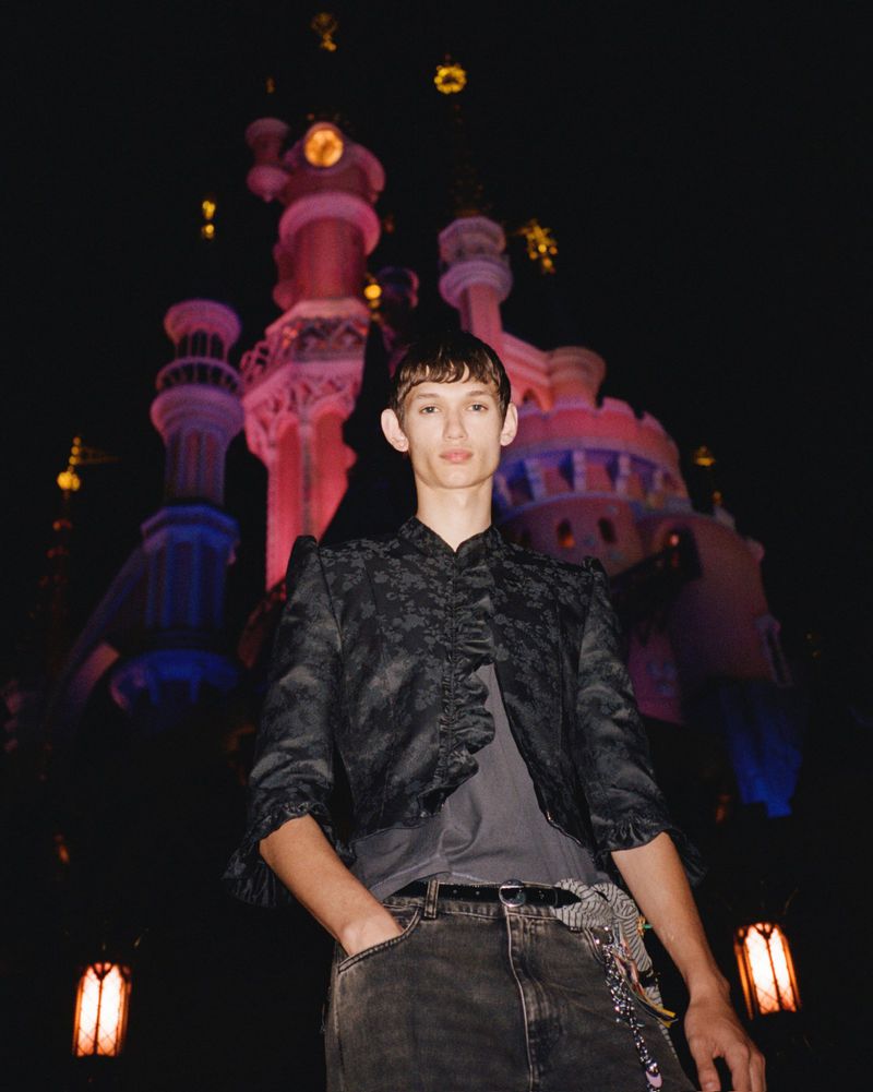 Young man in black jacket posing in front of a brightly illuminated castle at night, wearing jeans and a grey top.