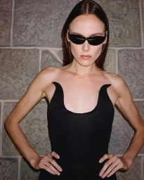 Model wearing dark sunglasses and a black dress standing against a stone wall background