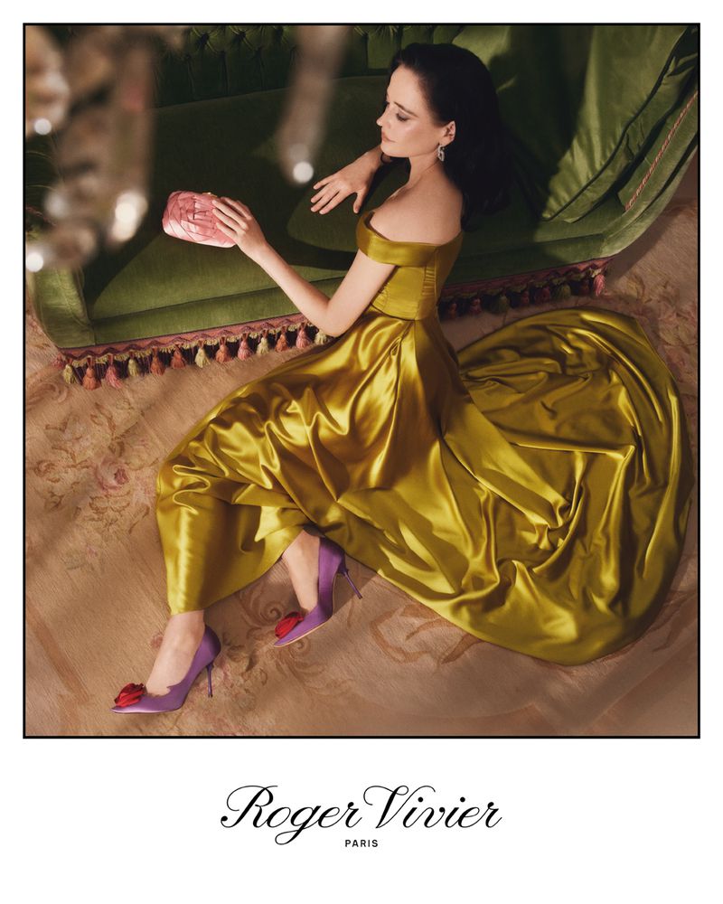 Woman in flowing yellow dress poses on green couch with pink clutch and purple heels in studio ad for Roger Vivier Paris.
