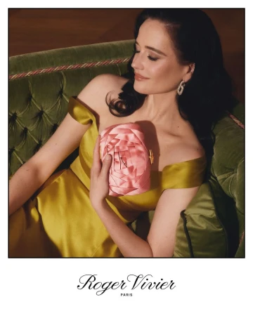 Eva Green poses with a pink floral clutch and shoulder-revealing yellow gown, seated on a green tufted chaise lounge in a studio setting.