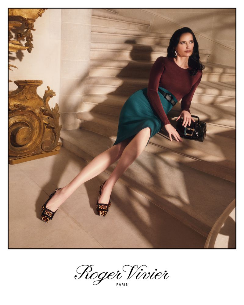 Elegant woman in long-sleeved maroon top and teal pencil skirt poses on stairs with a black purse and decorative shoes.