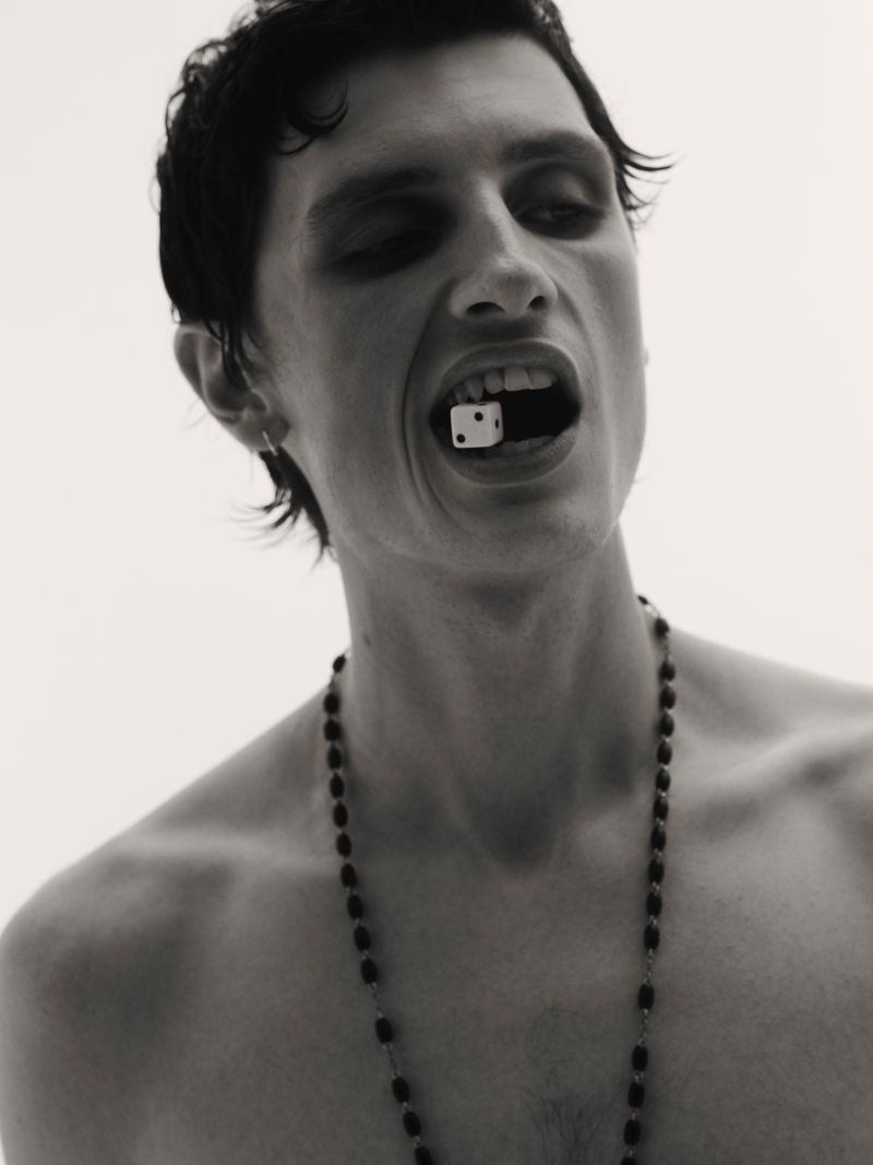 Portrait of a bare-chested adult male with dark hair holding a single die in his mouth and wearing a beaded necklace against a white background.