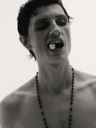 Portrait of a bare-chested adult male with dark hair holding a single die in his mouth and wearing a beaded necklace against a white background.