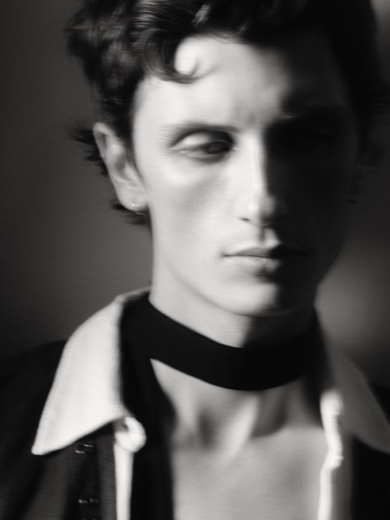 Monochrome portrait of a young adult male with dark, wavy hair wearing a jacket and black choker against a dark background.