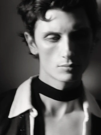 Monochrome portrait of a young adult male with dark, wavy hair wearing a jacket and black choker against a dark background.