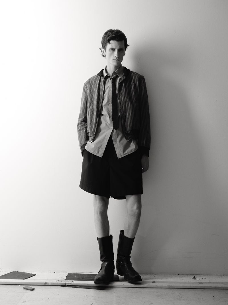 Black and white portrait of a young man in a shirt, jacket, shorts, and boots, standing against a simple wall in an indoor setting.