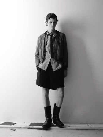 Black and white portrait of a young man in a shirt, jacket, shorts, and boots, standing against a simple wall in an indoor setting.