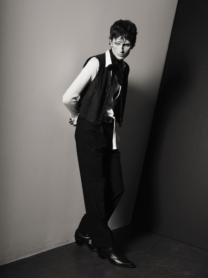 Monochrome fashion shot of a stylish adult male model in a vest, dress shirt, and slacks posing in a photo studio with a neutral backdrop.