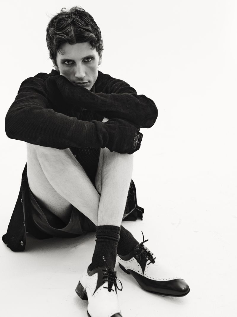 Monochrome portrait of a young adult man with dark wavy hair, wearing a black jacket, seated with black-and-white wingtip shoes on a white backdrop.