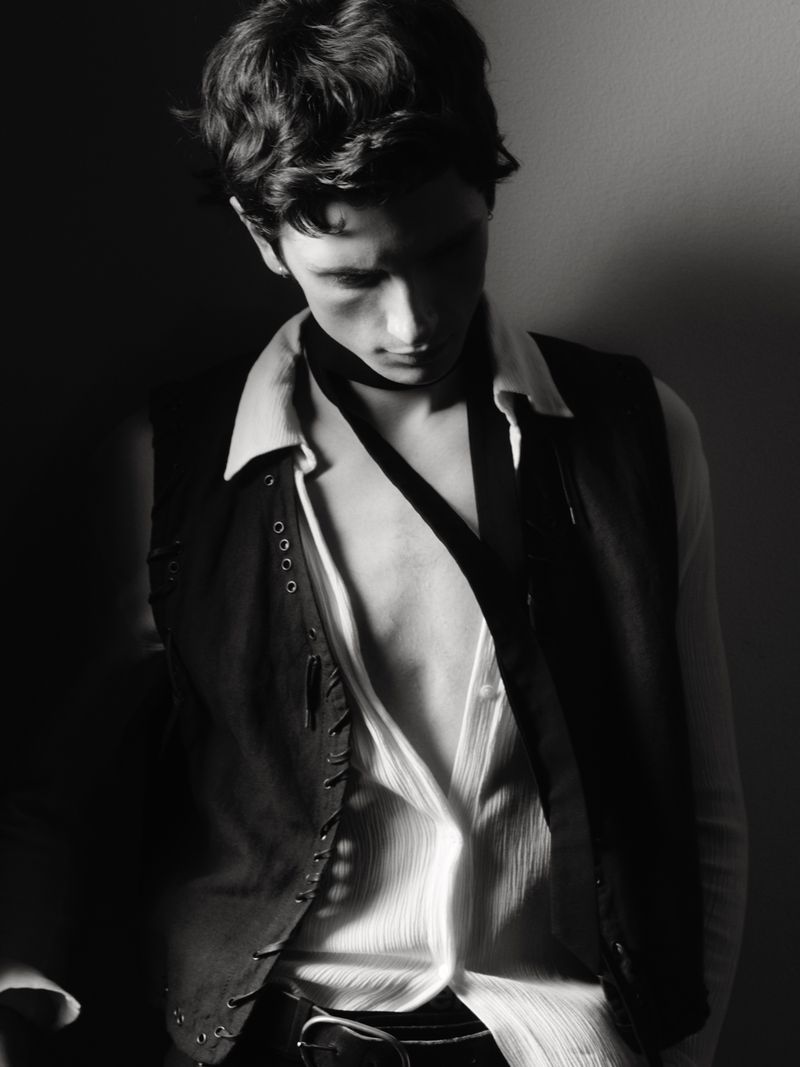 Black and white portrait of a young adult male in an unbuttoned shirt and studded vest looking down in an indoor studio setting.