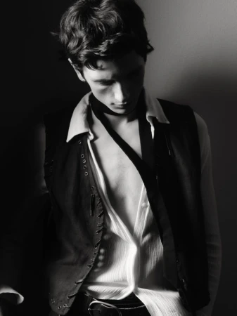 Black and white portrait of a young adult male in an unbuttoned shirt and studded vest looking down in an indoor studio setting.