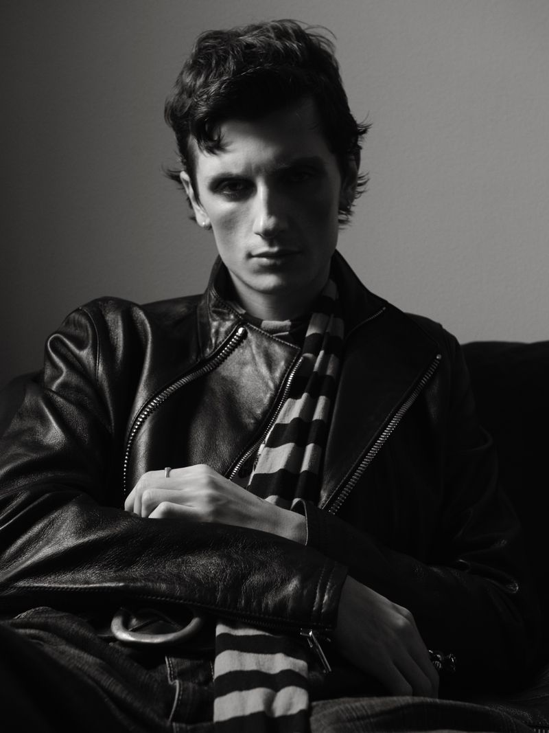 Monochrome portrait of a stylish adult male wearing a leather jacket and striped scarf, seated indoors with soft lighting and a neutral background.