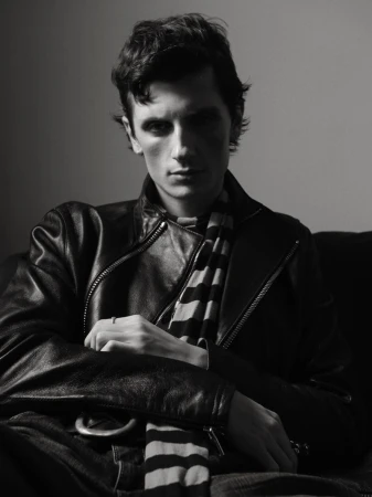 Monochrome portrait of a stylish adult male wearing a leather jacket and striped scarf, seated indoors with soft lighting and a neutral background.