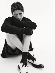 Monochrome portrait of a seated man wearing dark clothing and two-tone spectator shoes against a white studio background.