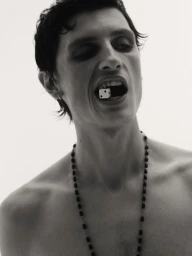 Black and white portrait of a shirtless man holding a white die in his mouth while wearing a dark beaded necklace against a plain backdrop.