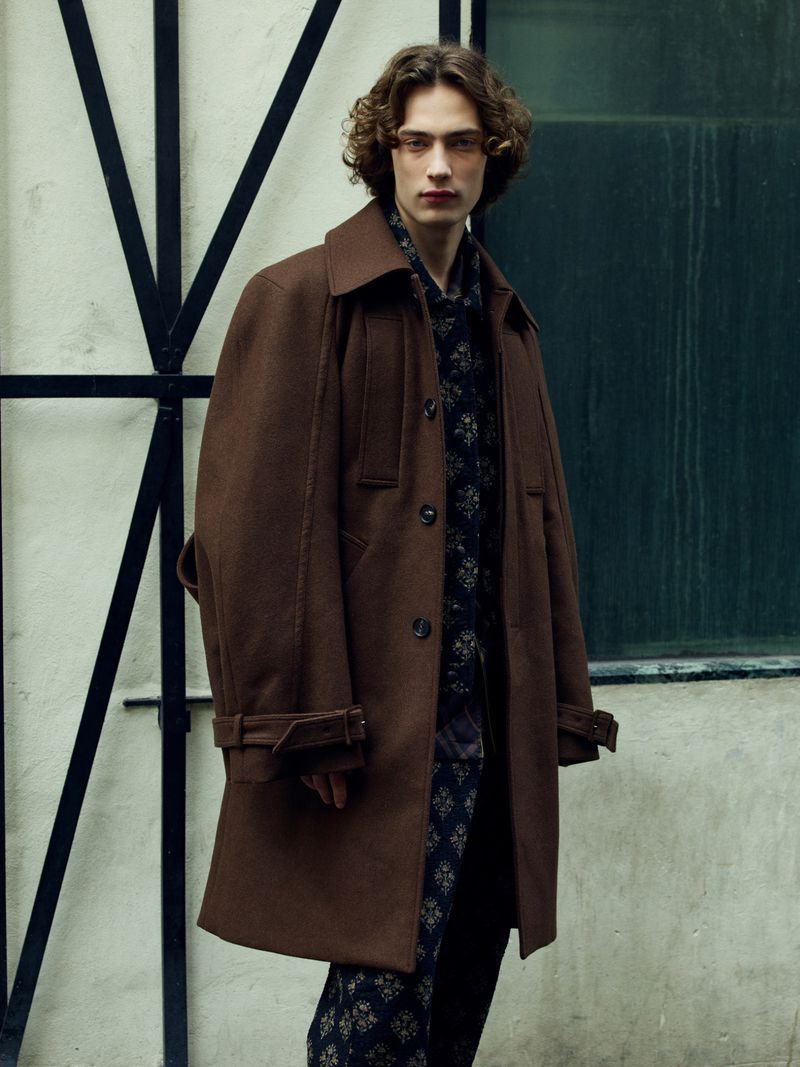 Model Freek Iven wearing a structured brown wool overcoat and a dark patterned suit against a pale wall with a black metal frame.