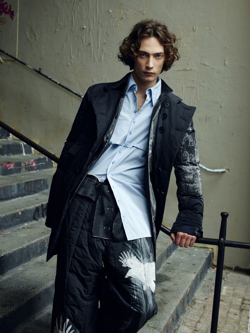 Fashionable young adult in a blue button-down shirt and dark coat and patterned pants poses on concrete steps with a metal railing.