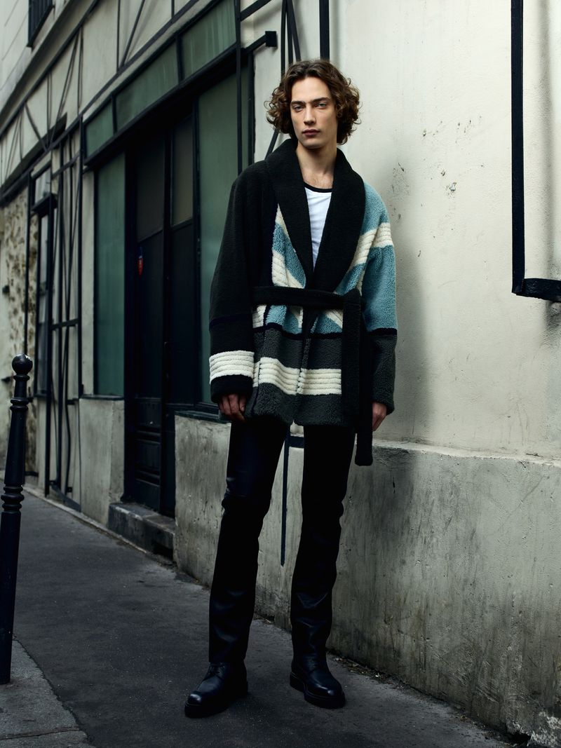 Male fashion model in stylish striped robe coat, black jeans, and boots poses in an urban setting with a neutral color palette.